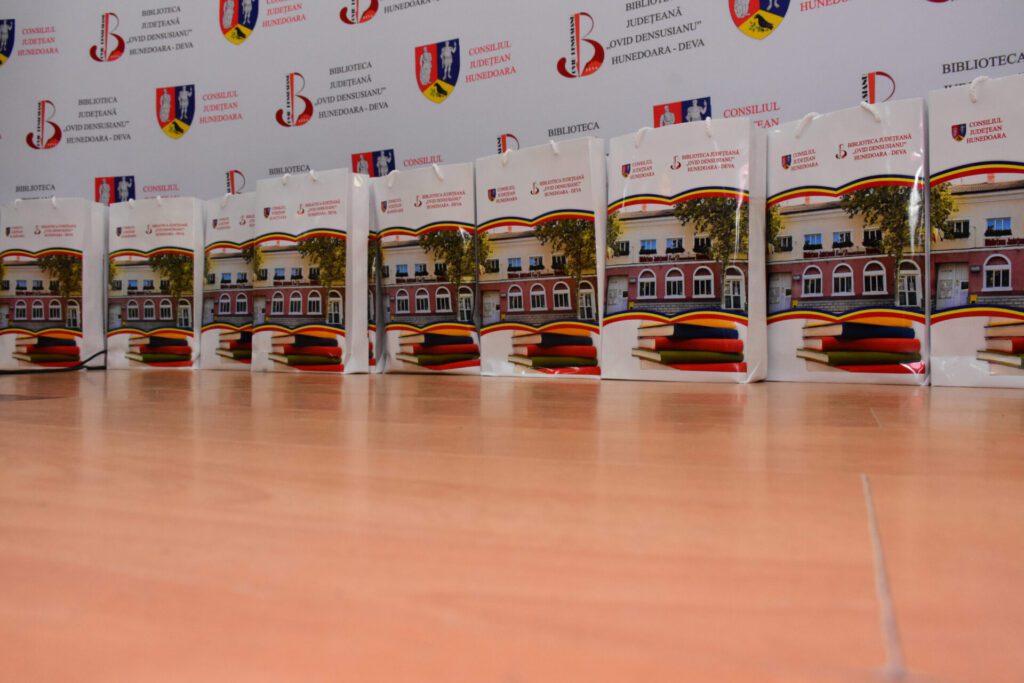 Pungi personalizate cu imaginea Bibliotecii Județene 'Ovid Densusianu' Hunedoara - Deva, așezate pe o suprafață de lemn, în cadrul unui eveniment.