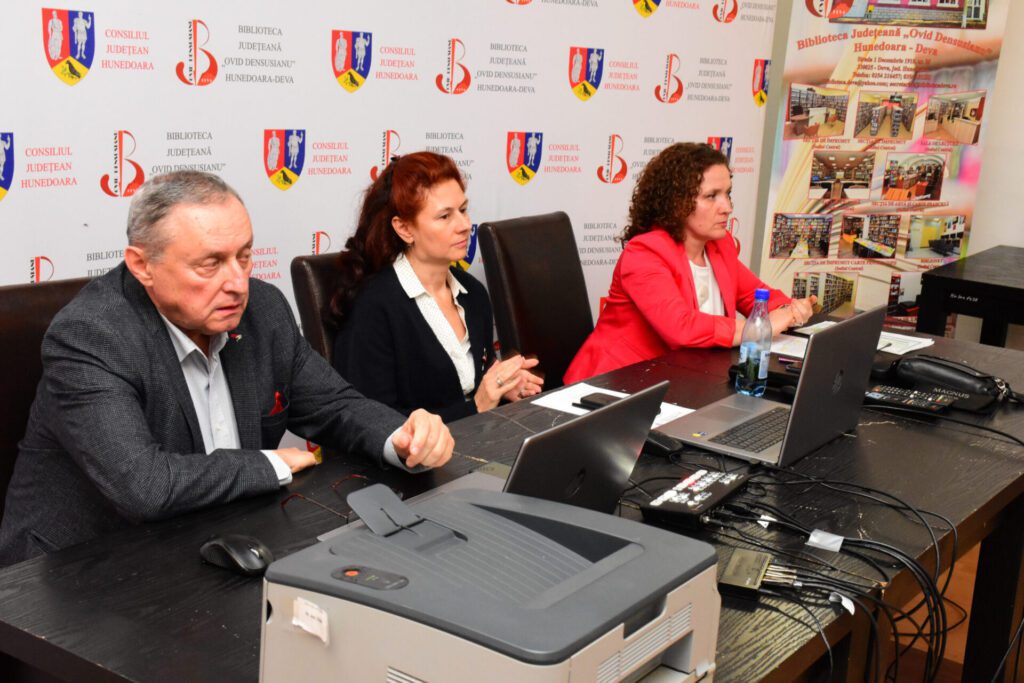 Patru persoane stau la o masă de lucru, discutând despre concursul de cultură generală organizat de Biblioteca Județeană Hunedoara - Deva. Trei dintre ele sunt femei, iar una este bărbat, toți având laptopuri și materiale de lucru pe masă. Fundalul afișează siglele instituțiilor implicate în organizare.