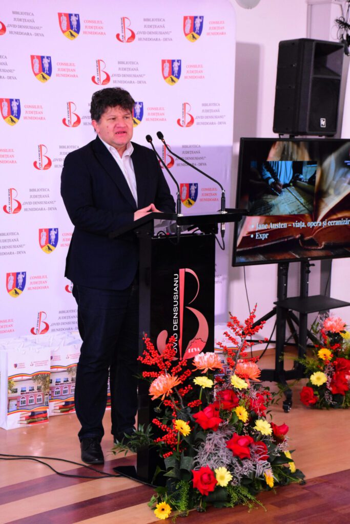 Un vorbitor la un eveniment organizat de Biblioteca Județeană „Ovid Densusianu” Hunedoara - Deva, stând în fața unui pupitru de speaker, cu flori decorative în prim-plan.