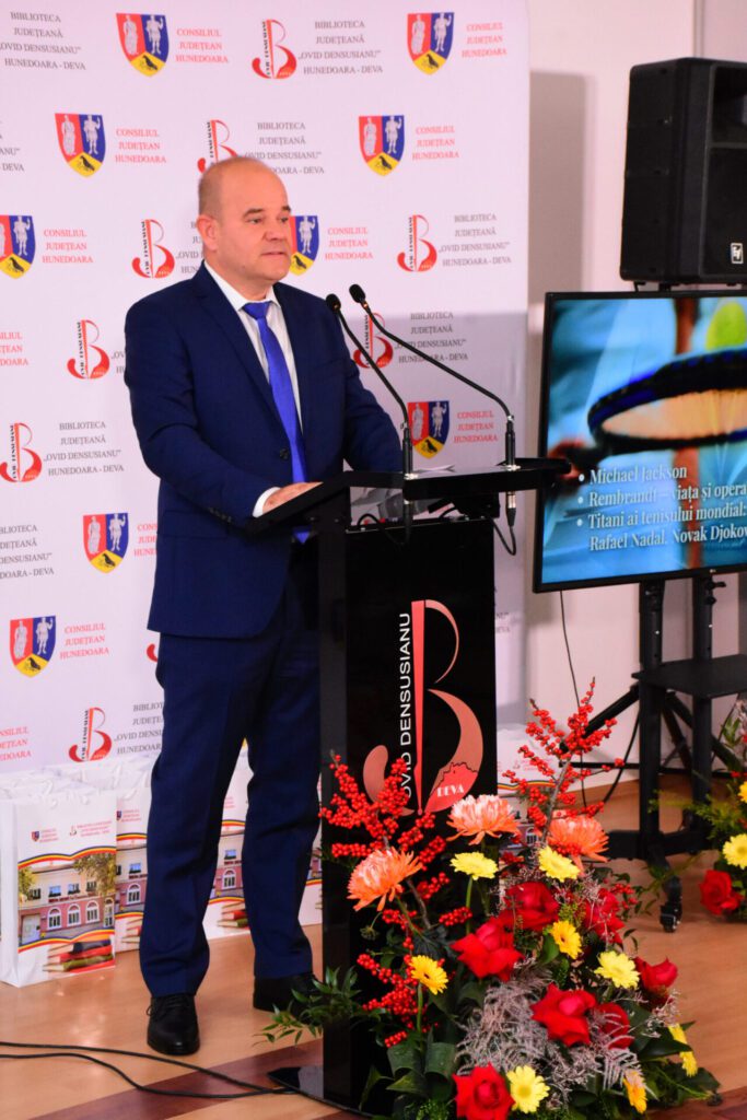 Un bărbat îmbrăcat în costum albastru, la un pupitru, ținând un discurs în fața unui public, cu un ecran în fundal care afișează informații despre concurenți și premii. Decor floral colorat pe masă.