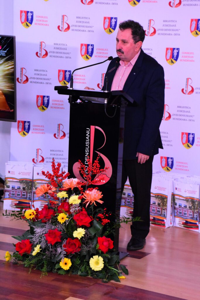 Un bărbat susține un discurs la un eveniment organizat de Biblioteca Județeană "Ovid Densusianu" Hunedoara - Deva, în fața unui panou cu siglele instituțiilor partenere și flori decorative.