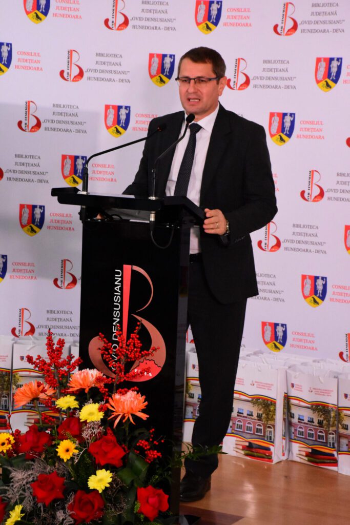 Un bărbat în costume formal vorbește la un pupitru, cu un aranjament floral colorat în față, în cadrul unei ceremonii organizate de Biblioteca Județeană „Ovid Densusianu” Hunedoara - Deva.