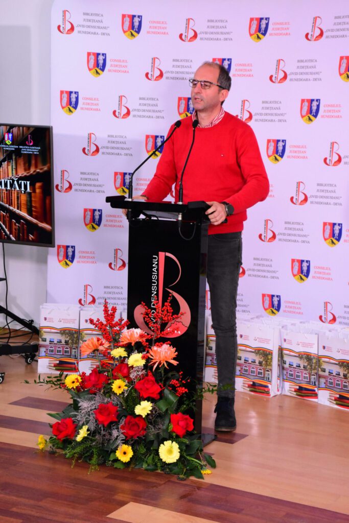 Un vorbitor angajat pe un podium, ținând un discurs, în fața unui banner al Bibliotecii Județene „Ovid Densusianu” Hunedoara - Deva, decorat cu flori colorate.