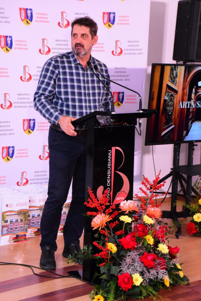 Un bărbat stă la un podium de discurs, în fața unui banner cu sigla Bibliotecii Județene "Ovid Densusianu" Hunedoara - Deva, înconjurat de flori colorate.