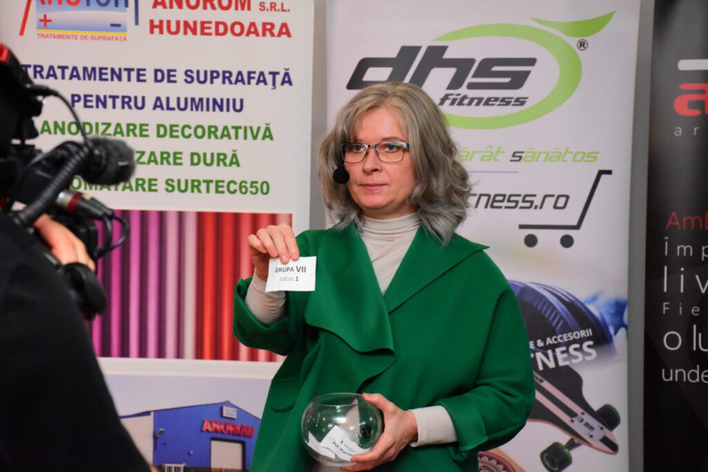 O persoană îmbrăcată într-o haină verde ține un bilet cu inscripția 'GRUPA VII LOCUL 1' în timp ce este filmată.