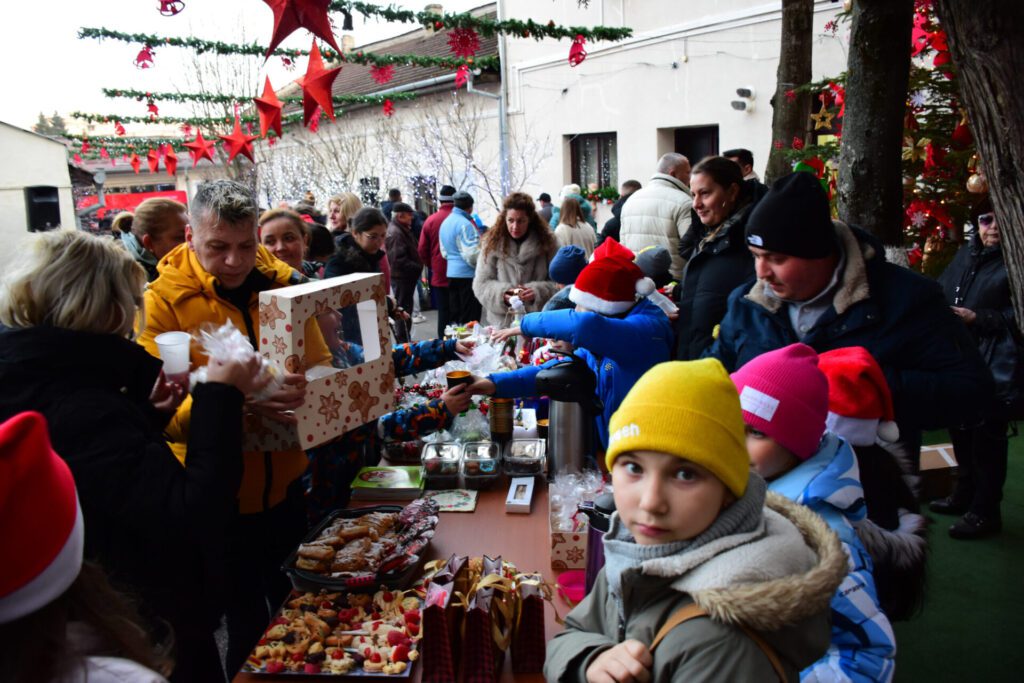 Oameni adunați în curtea bibliotecii, participând la un târg de Crăciun cu mese pline de produse artizanale și decoruri festive.
