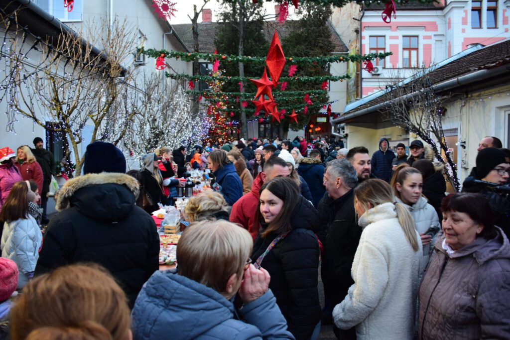 Oameni adunați într-o curte decorată festiv, cu lumini de Crăciun și ornamente, unde se vând produse handmade de sărbători.