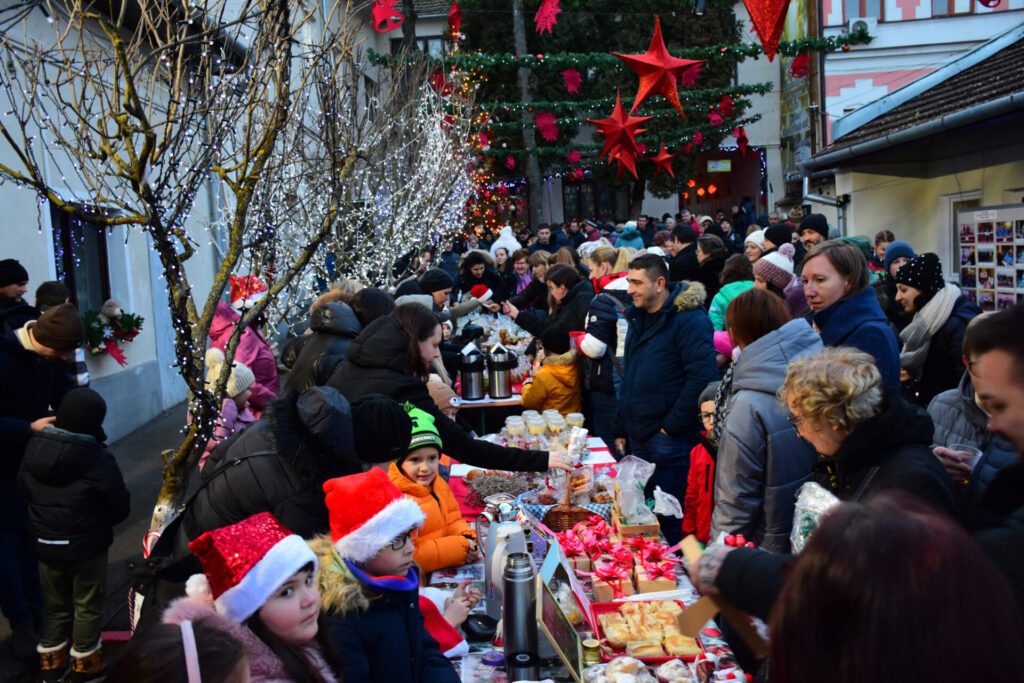 Scenă din curtea unei biblioteci, plină de oameni care admirează și cumpără obiecte decorative de Crăciun, înconjurați de nuanțe festive de lumină și decorațiuni.
