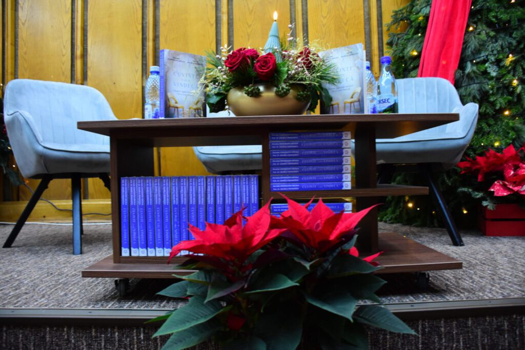 Mesea decorată cu flori roșii și cărți pe o masă, în fața unor scaune, cu un fundal festiv de Crăciun.