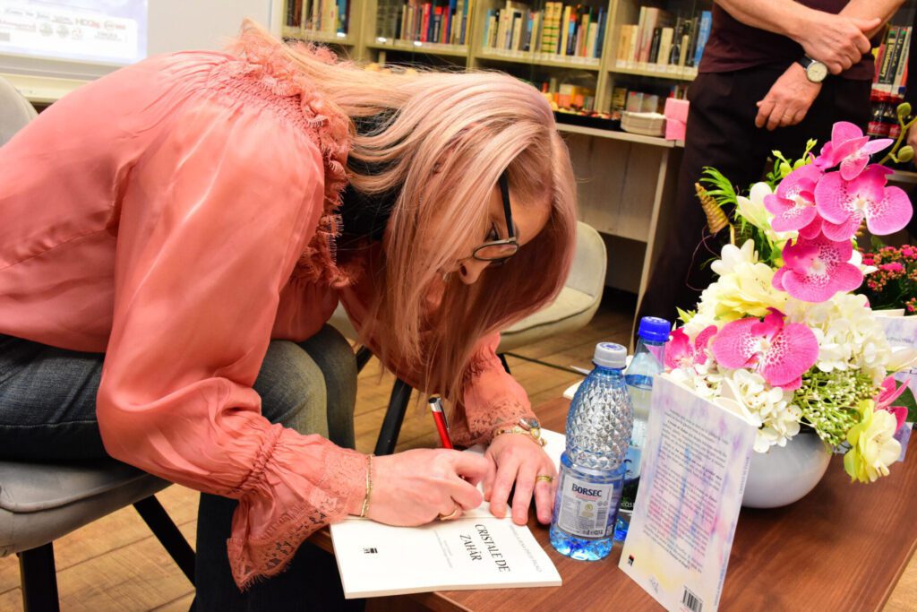 O persoană semnează o carte, având un buchet de flori și sticle de apă pe masă. Aceasta poartă o bluză roz și ochelari, fiind concentrată asupra scrisului.