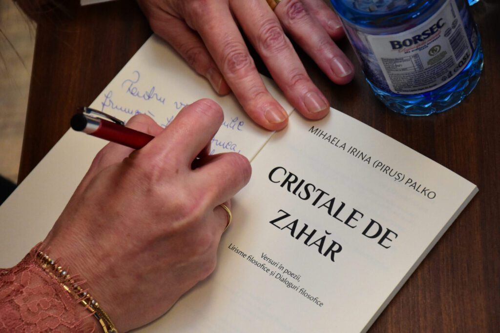 O persoană scrie un mesaj într-o carte intitulată 'Cristale de Zahăr', având un pix roșu în mână, lângă o sticlă de apă.